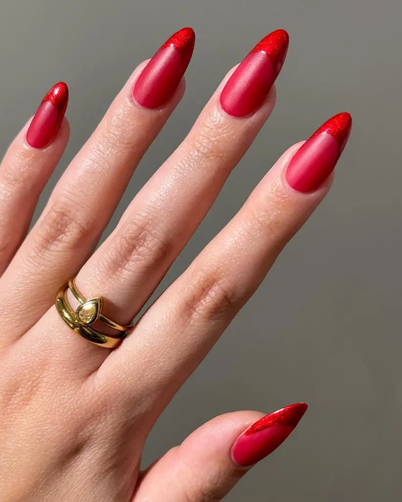 Glossy red almond-shaped nails with a sparkling finish, highlighted by natural lighting and adorned with a single gold band ring
