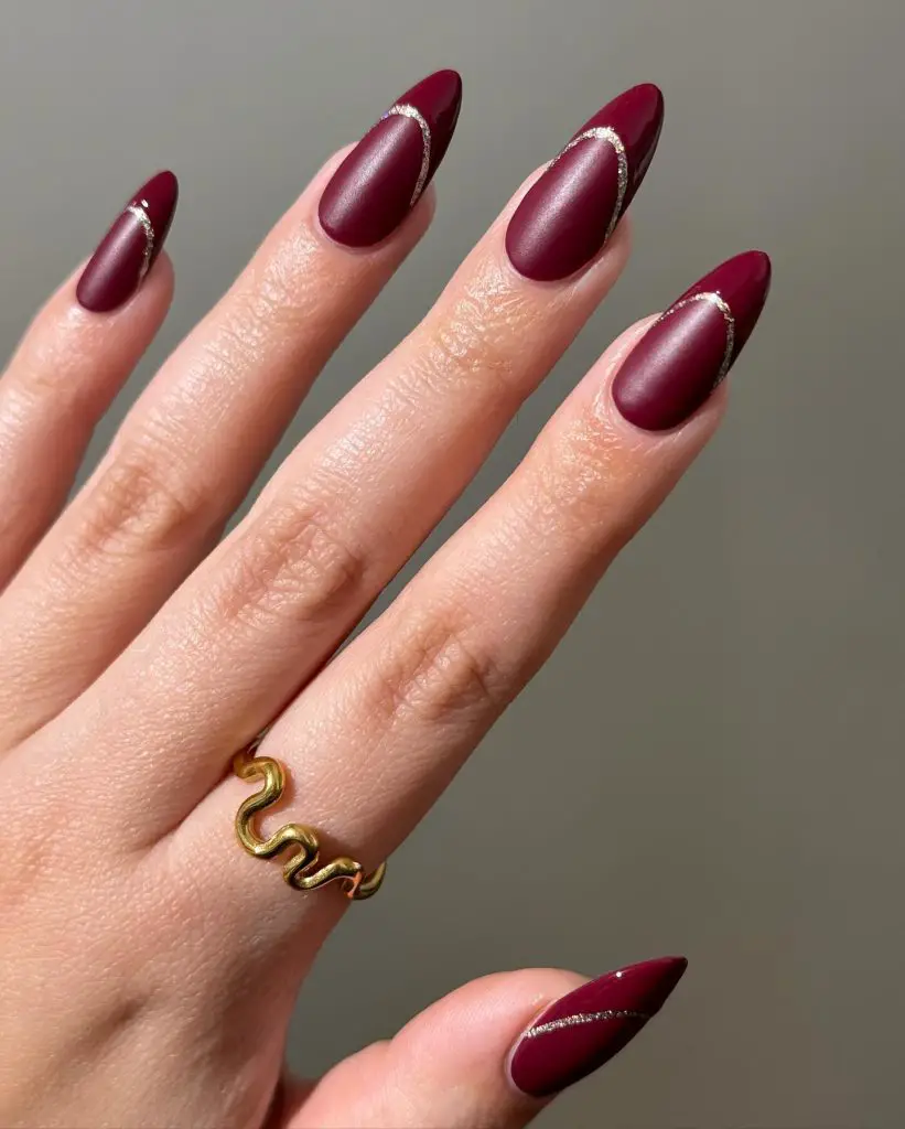 Matte burgundy stiletto nails featuring a single horizontal stripe of silver glitter on each ring finger, complemented by a wavy gold ring