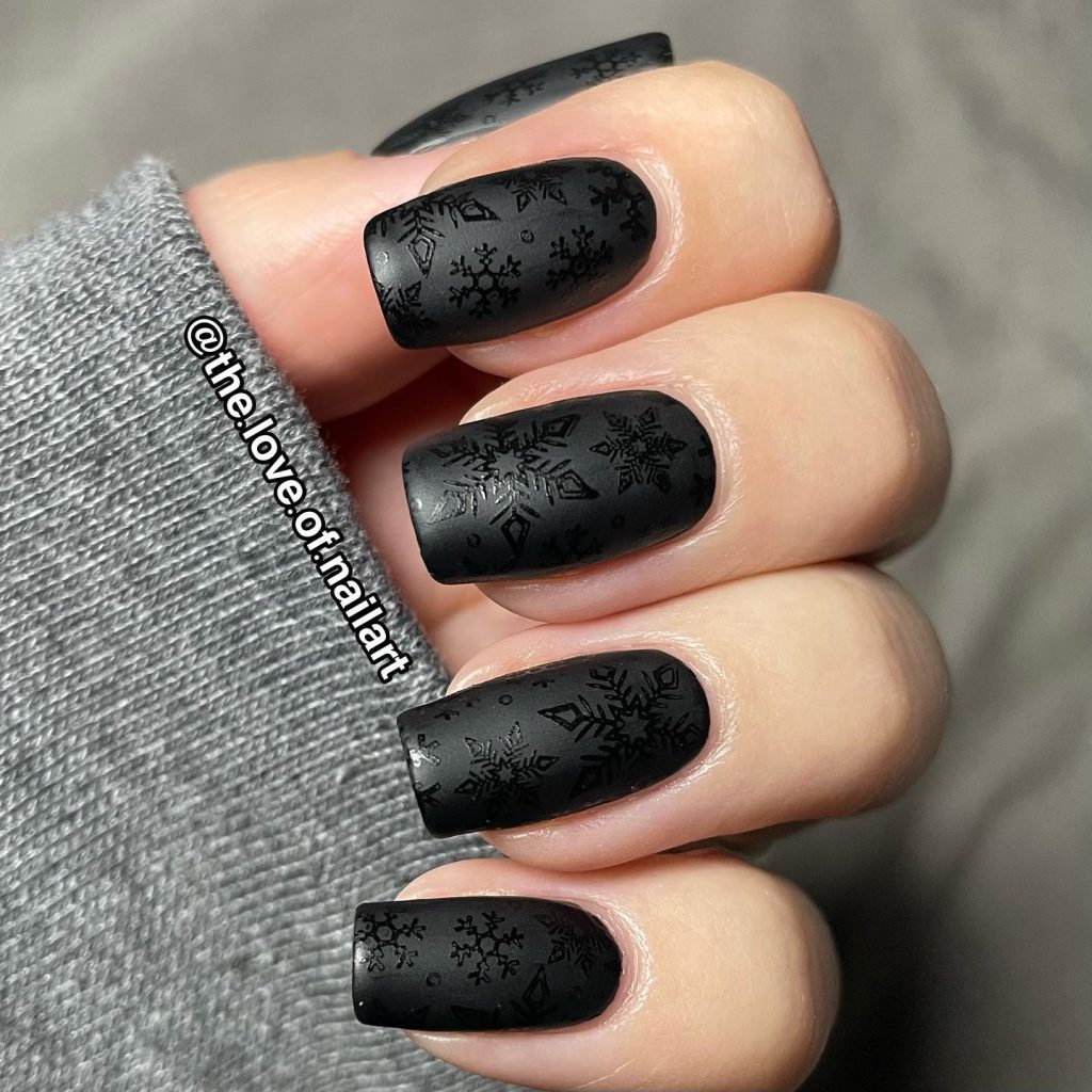 Matte black nails with a subtle black snowflake stamp design, set against a soft grey fabric background.